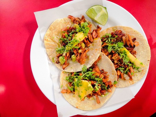 Un plato que muestra tres tacos de pastor acompañados con salsa, limon y piña